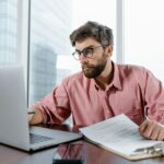 A businessman in an office reviews documents and data on a laptop, concentrating on financial analysis.